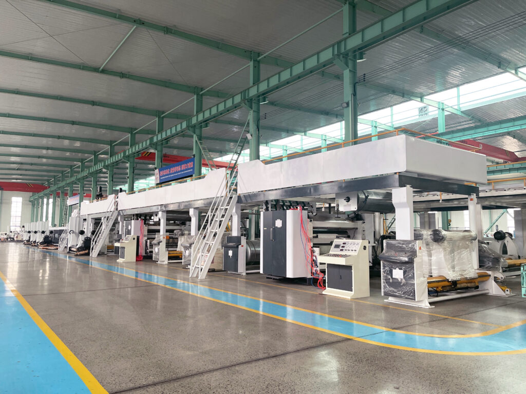 A large-scale corrugated cardboard making machine line stands on a concrete floor inside a vast industrial warehouse. The image shows multiple connected machine units with white bodies and a control console, along with metal stairs leading to an upper level. The green steel structure of the warehouse is visible overhead. This high-efficiency corrugated cardboard making machine is designed for the high-volume production of durable packaging materials.